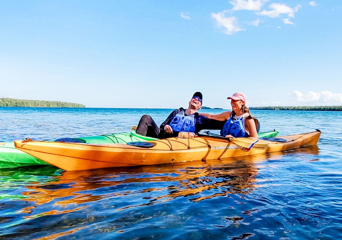 A man and a woman sitting in solo sea kayaks are laughing while taking a break during a kayak tour with Woods & Waters in Lake Huron's Les Cheneaux Islands.