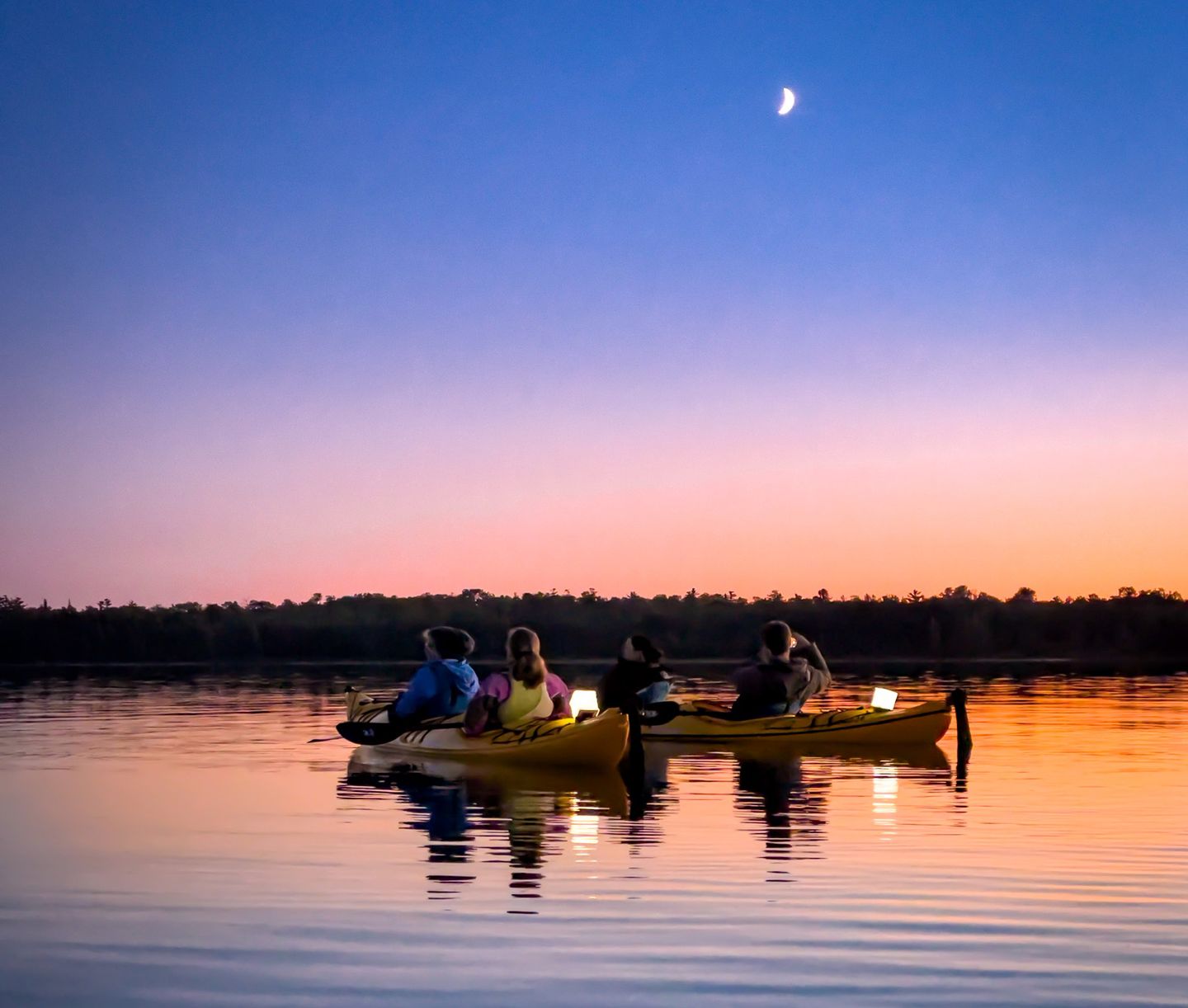 Two tandem kayaks with illuminated solar lanterns sit on calm waters at dusk in Lake Huron's Les Cheneaux islands while on a sunset kayak tour with Woods & Waters. A crescent moon is visible in the sky above them.