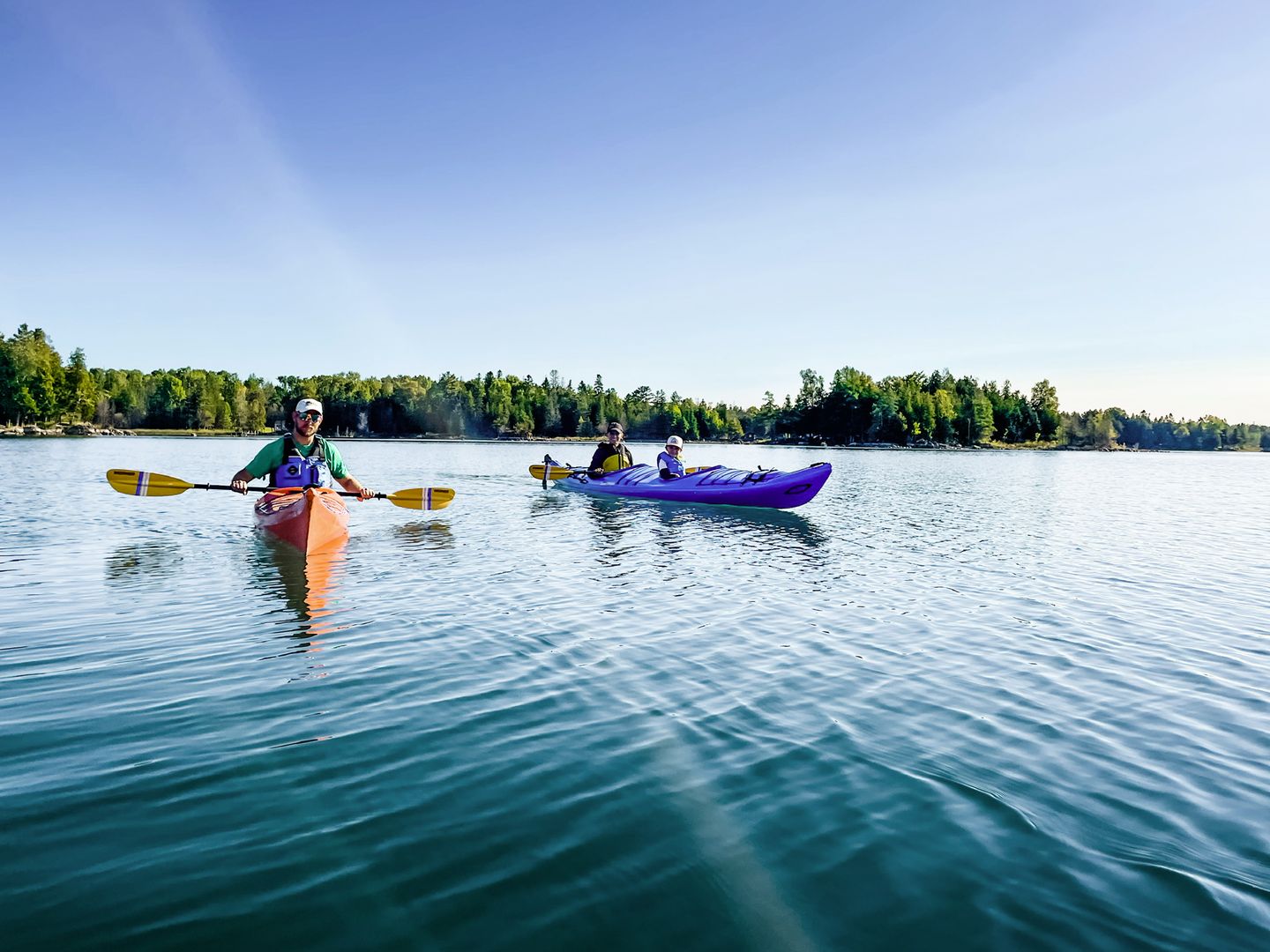 Two kayaks, an orange solo kayak, and a blue tandem kayak with a parent and child, take a quick break for the camera while on a Little Dipper kayak tour in Lake Huron's Les Cheneaux Island.