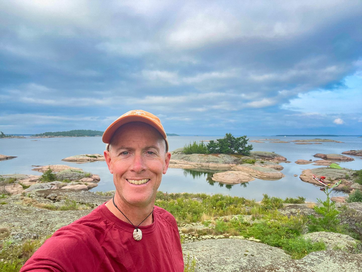 A middle-aged man in a red shirt and baseball cap stands on the shoreline with small rocky island behind him and a red kayak on the far right.