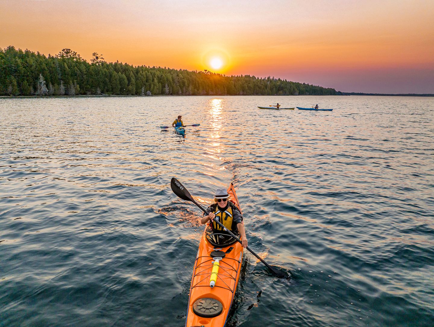 Several solo sea kayaks are paddling toward the camera on Wilderness Bay in Lake Huron's Les Cheneaux Islands while on a sunset kayak tour with Woods & Waters. The sun is setting behind an island.