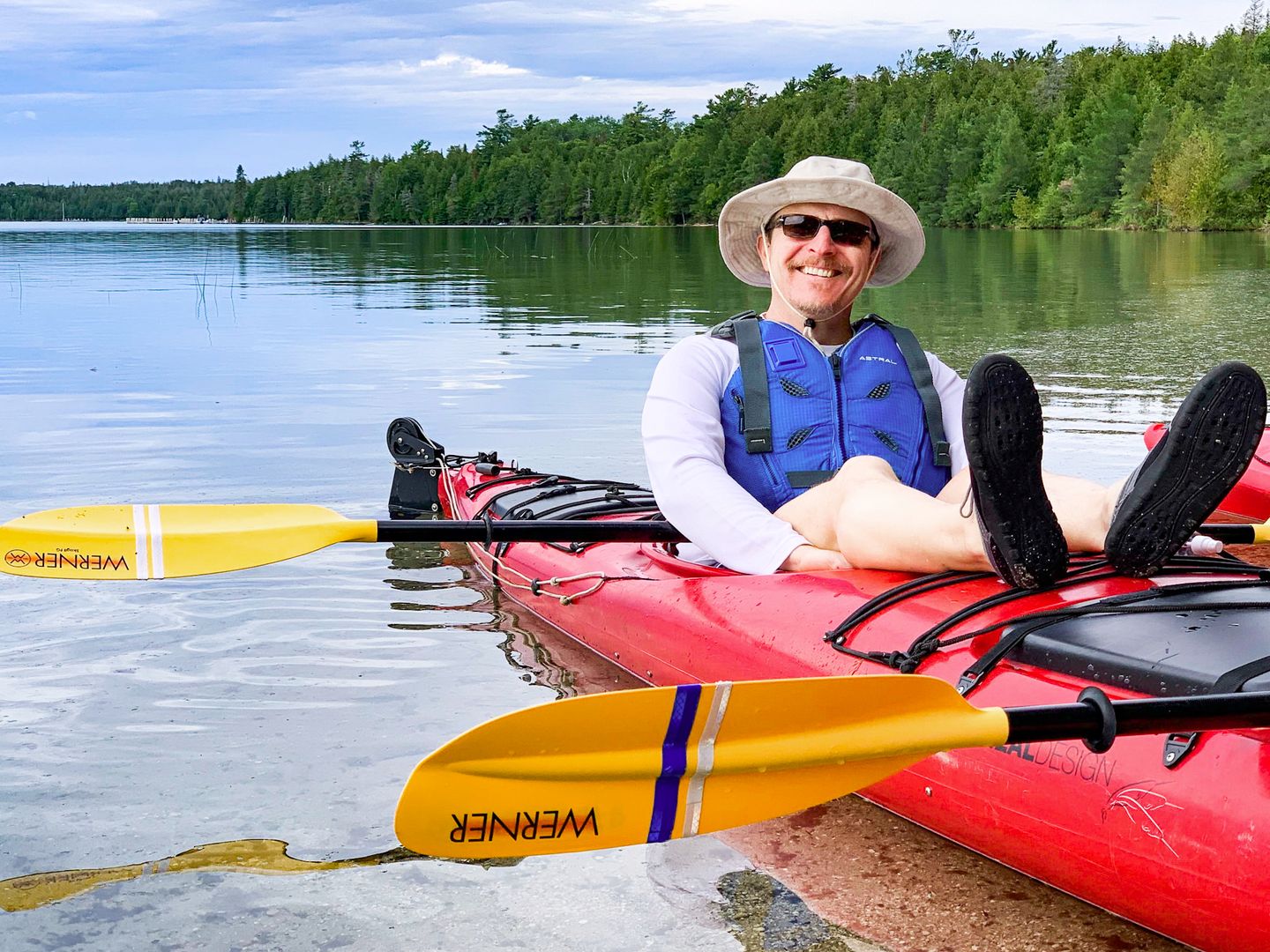 A man sits in the rear cockpit of a tandem seas kayak with his feet propped up on the deck and is smiling at the camera. He is taking a break in Marquette Bay in Lake Huron's Les Cheneaux Islands while on a Bigger Dipper kayak tour with Woods & Waters. 