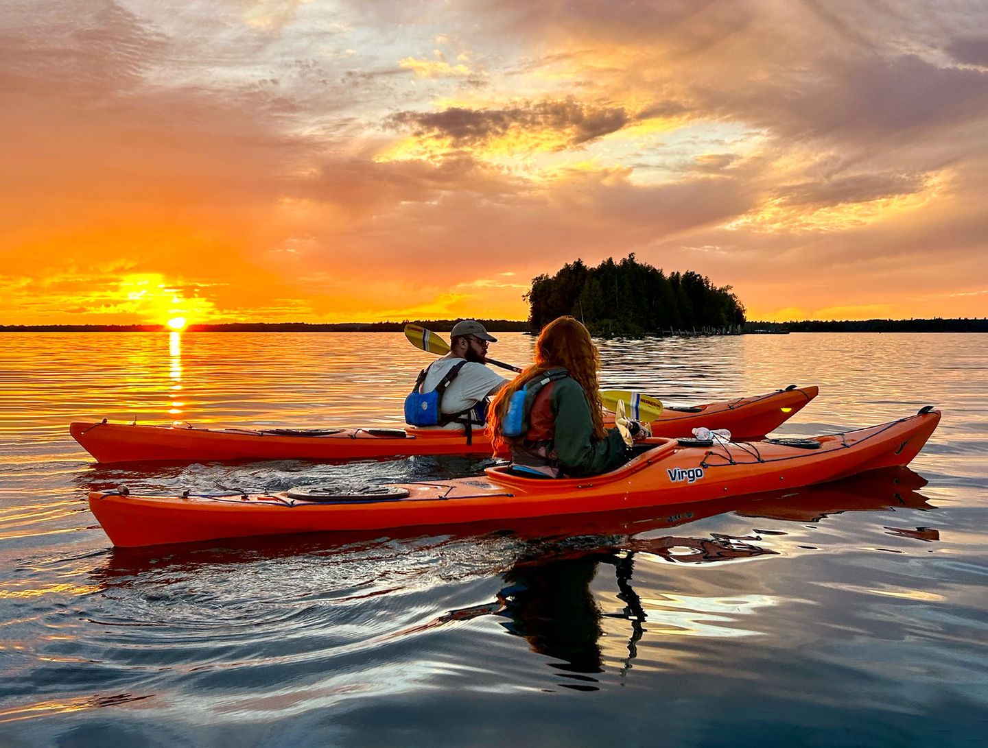 A man and a woman, each in a solo sea kayak, paddle on calm the waters of Lake Huron in the Les Cheneaux Islands with a beautiful sunset and clouds beyond them.
