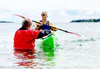 A teen-aged boy squinting from splashing water practices paddle bracing in a sea kayak while a man in a red dry suit assists.