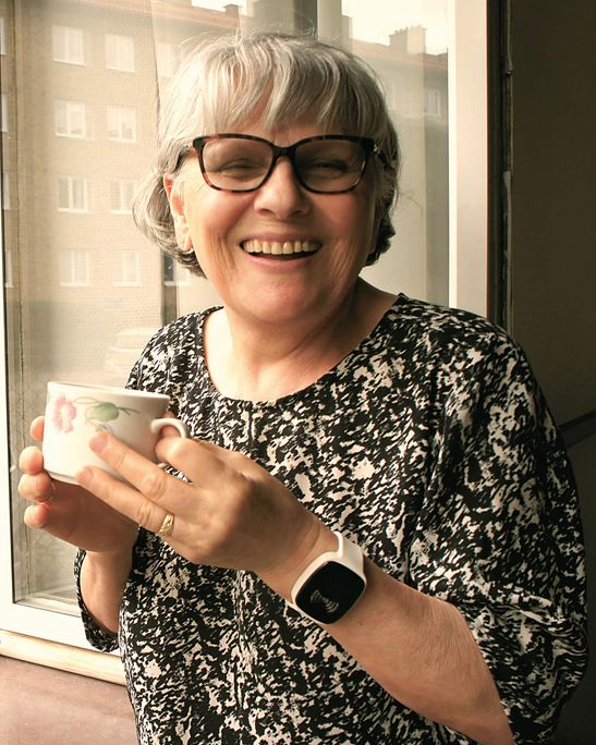 Leende äldre kvinna står vid ett öppet fönster med en kopp kaffe i handen. Hon bär TM-ALERT i vitt armband på vänster handled, en modul som vibrerar vid notiser. Grönskande innergård och flerbostadshus syns i bakgrunden.